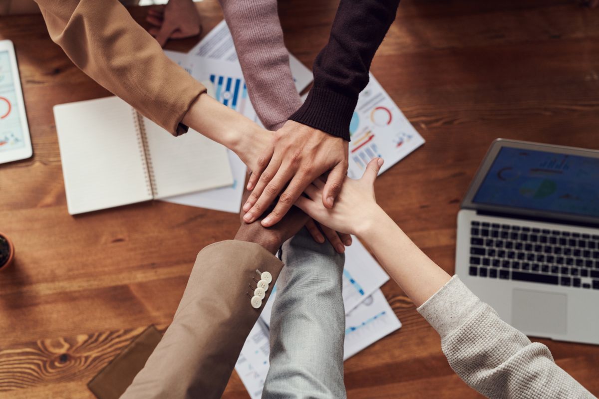 Three people stacking their hands together in a team huddle over a desk covered with printed charts and a laptop — illustrating collaborative portfolio project work