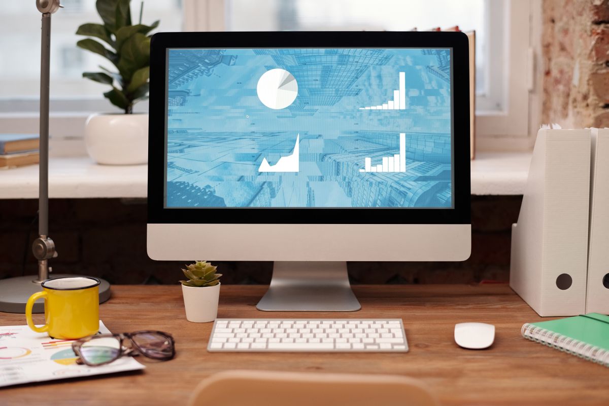 A desk setup with a desktop monitor showing bar charts, pie charts, and line graphs in blue, surrounded by folders, reports, glasses, and a keyboard — illustrating how a recruiter scans data analyst work samples