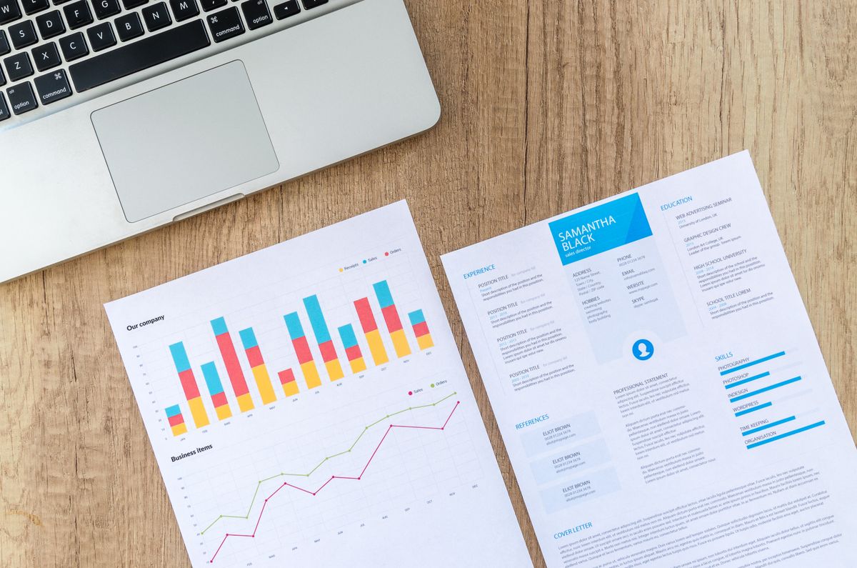 A wooden desk with a laptop and a printed resume next to printed bar chart reports — the visual setup of a job seeker reviewing profile materials